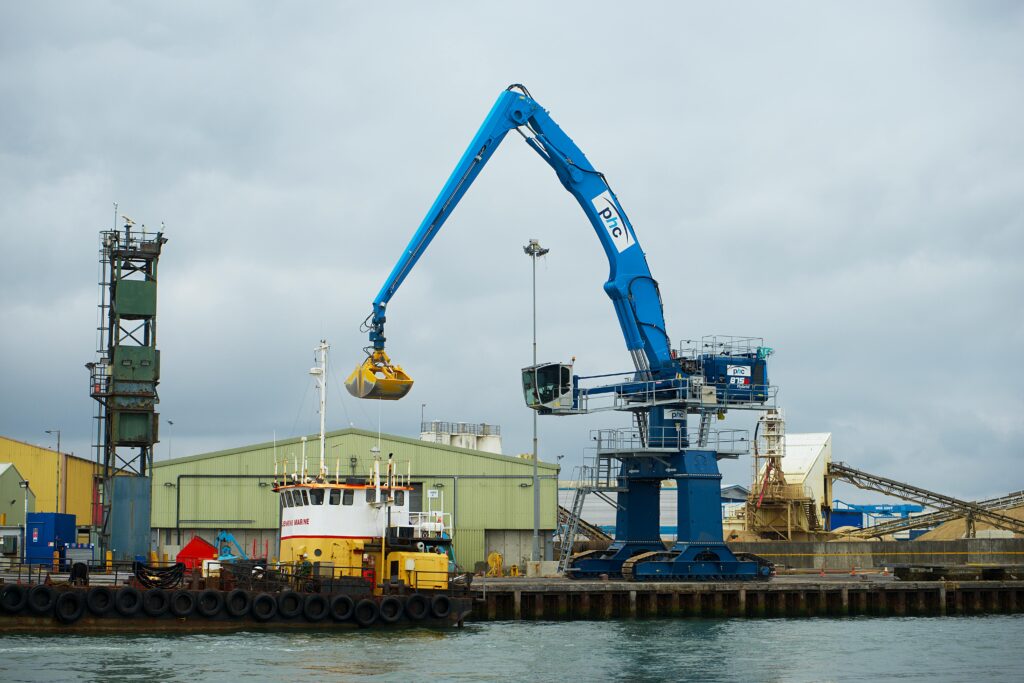 A large blue crane loading a vessel at Poole Harbor, England, surrounded by warehouses and industrial structures.